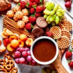 A dessert board with fruit, waffles, pretzels, and with a bowl of fondue in the center.