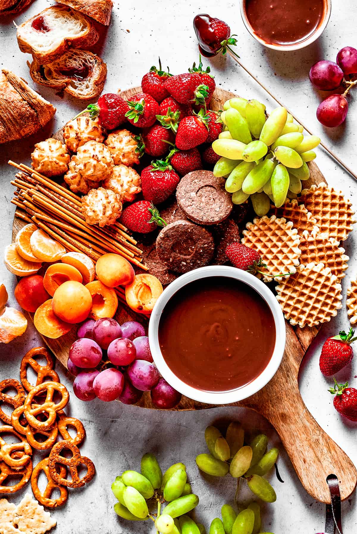 A dessert board with fruit, waffles, pretzels, and with a bowl of fondue in the center.