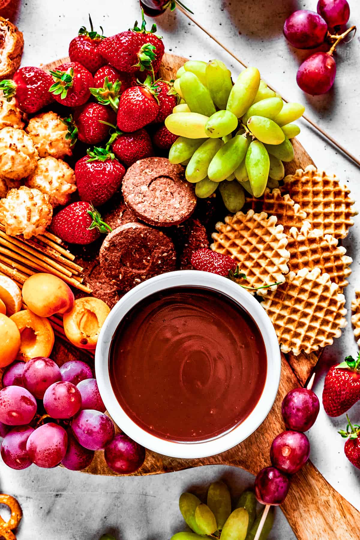 A dessert board with fruit, waffles, pretzels, and a bowl of fondue in the center.
