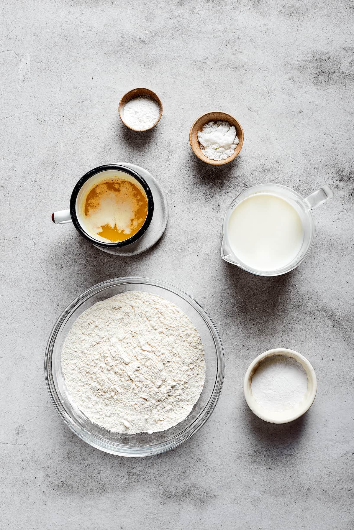 Overhead view of the ingredients needed for butter swim biscuits: a bowl of flour, a cup of melted butter, a pyrex of buttermilk, a bowl of sugar, a bowl of salt, and a bowl of baking powder
