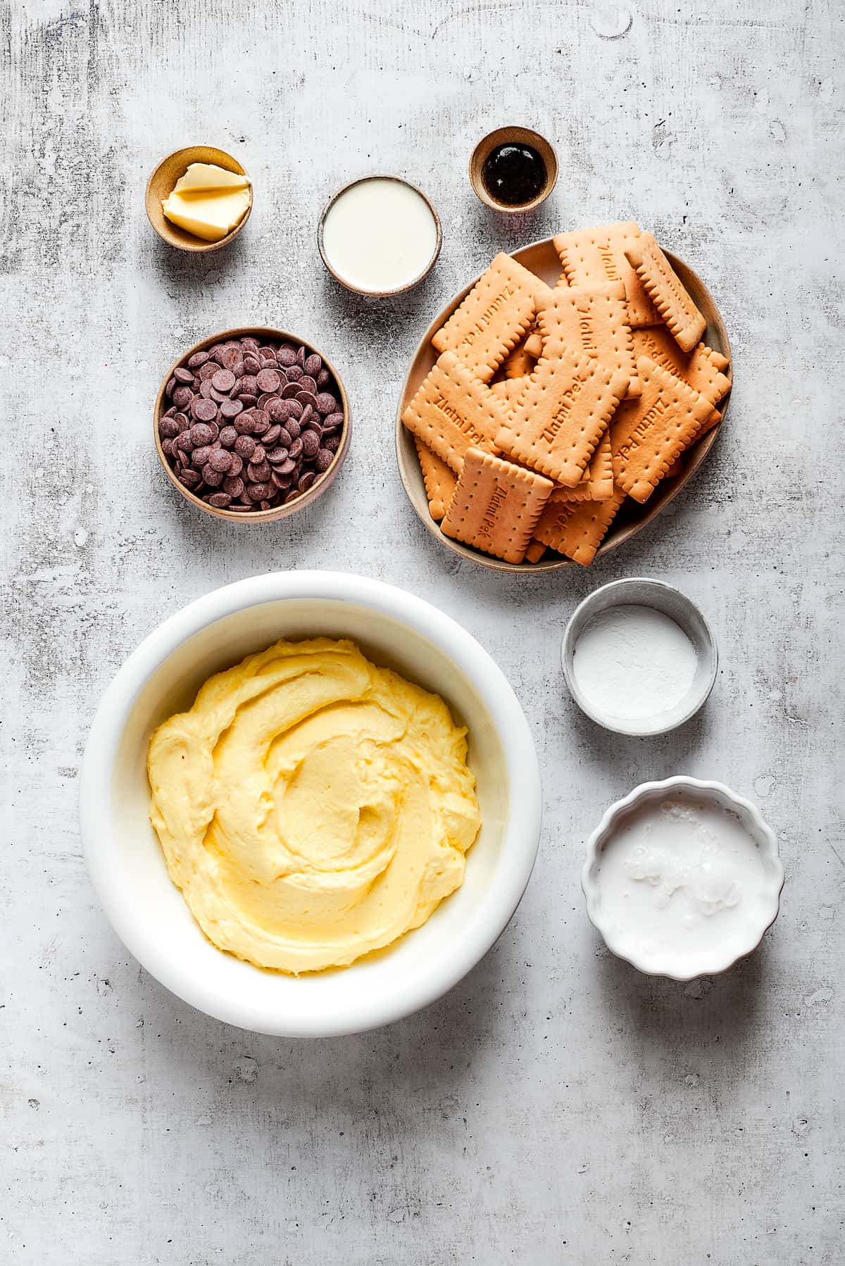 Overhead view of the ingredients needed for chocolate eclair cake: a bowl of vanilla pudding, a bowl of cookies, a bowl of chocolate chips, a bowl of cream, a bowl of milk, a bowl of butter, a bowl of vanilla, and a bowl of powdered sugar.