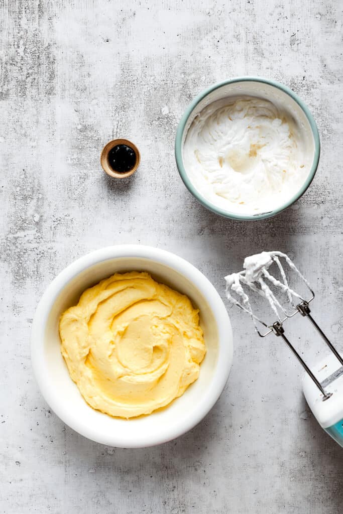 A bowl of vanilla pudding next to a bowl of whipped cream and a bowl of vanilla extract, with a beater.