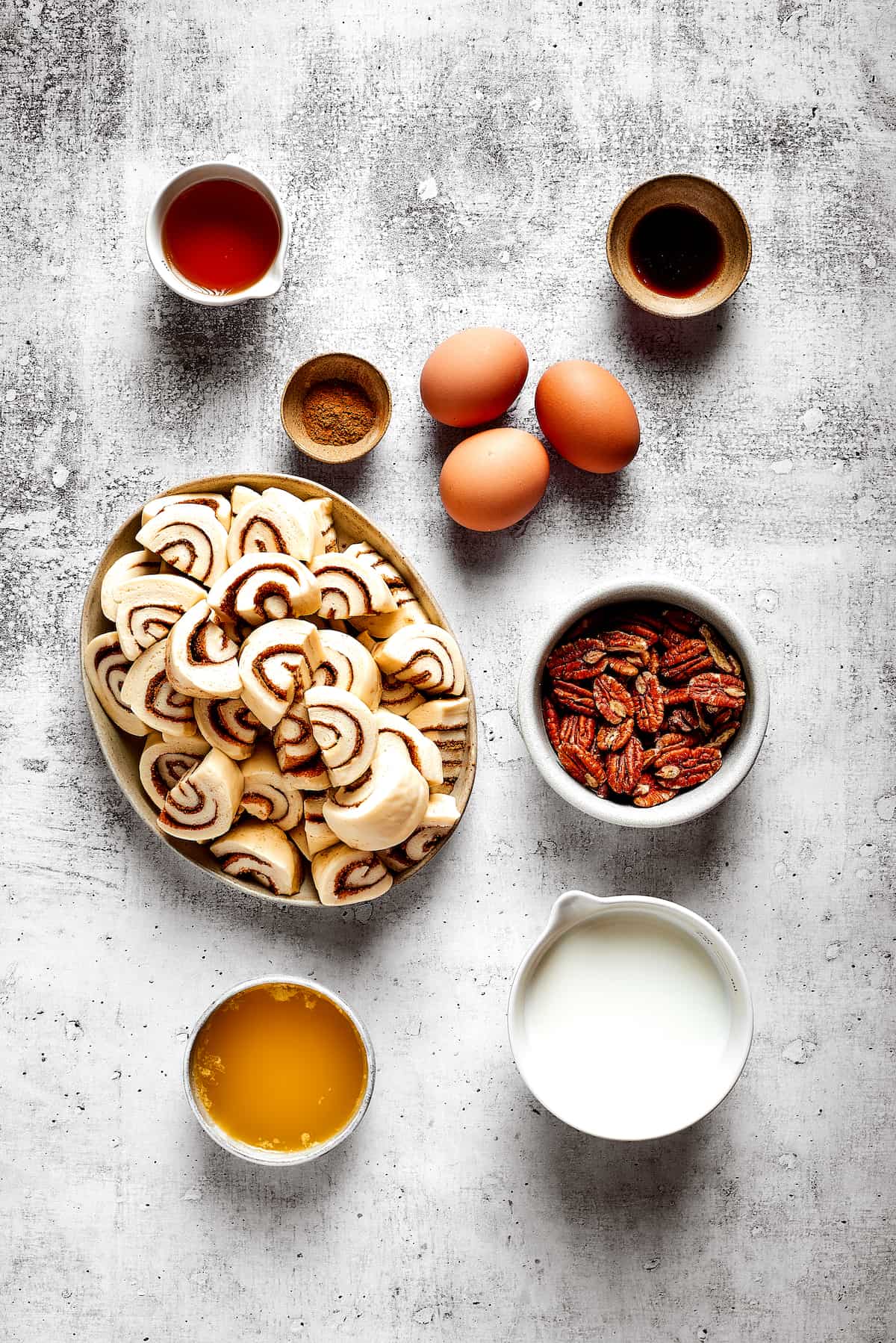Overhead view of the ingredients needed for cinnamon roll casserole: a plate of cinnamon roll pieces, a bowl of milk, a bowl of maple syrup, a bowl of pecans, three eggs, a bowl of cinnamon, a bowl of vanilla, and a bowl of melted butter