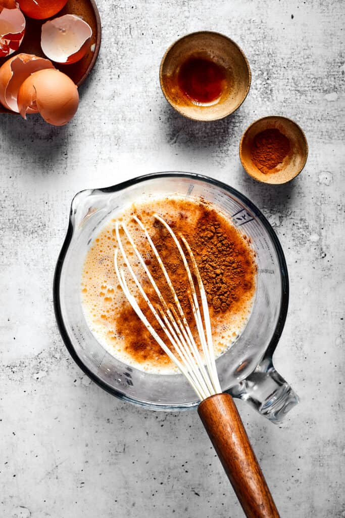 A pyrex filled with eggs and milk beat together with cinnamon on top, with a whisk, next to some bowls and egg shells