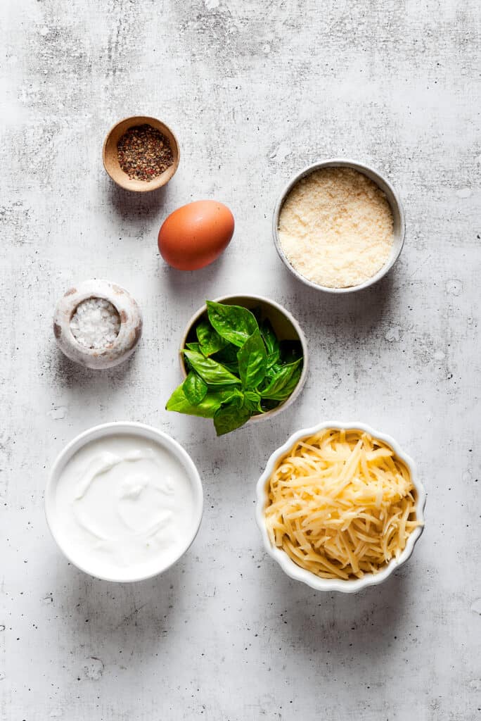 Overhead view of the ingredients needed for cheese filling: a bowl of parmesan, a bowl of mozzarella, a container of ricotta, a bowl of basil, an egg, a bowl of salt, and a bowl of pepper.