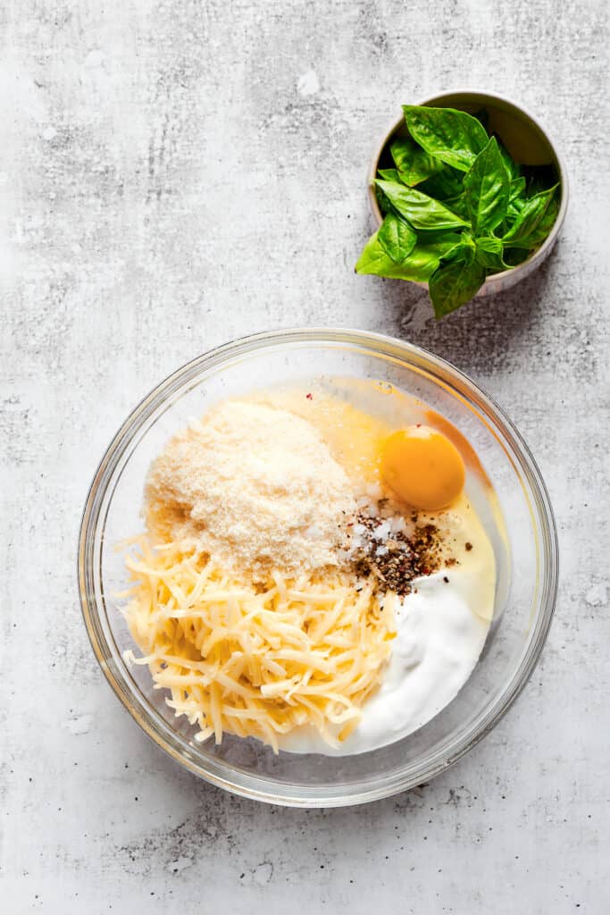 Overhead view of a mixing bowl filled with parmesan cheese, mozzarella cheese, ricotta, an egg, salt, and pepper, next to a bowl of basil.
