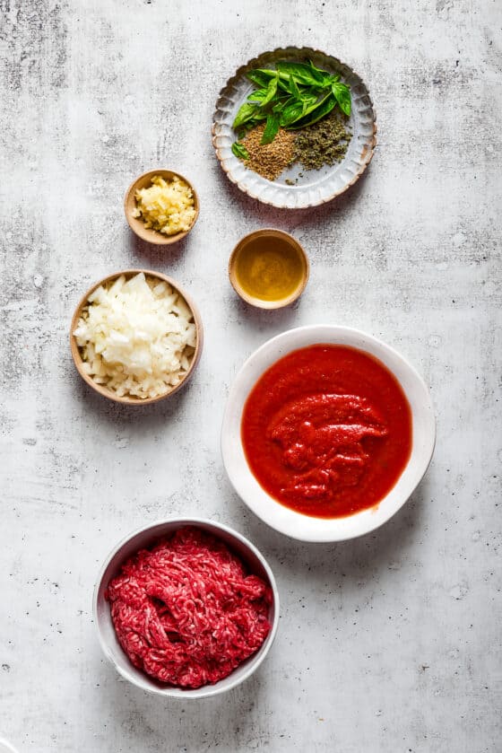 Overhead view of the ingredients needed for meat sauce: a bowl of ground beef, a bowl of marinara sauce, a bowl of chopped onion, a bowl of minced garlic, a bowl of olive oil, and a plate with fresh basil and dried herbs.