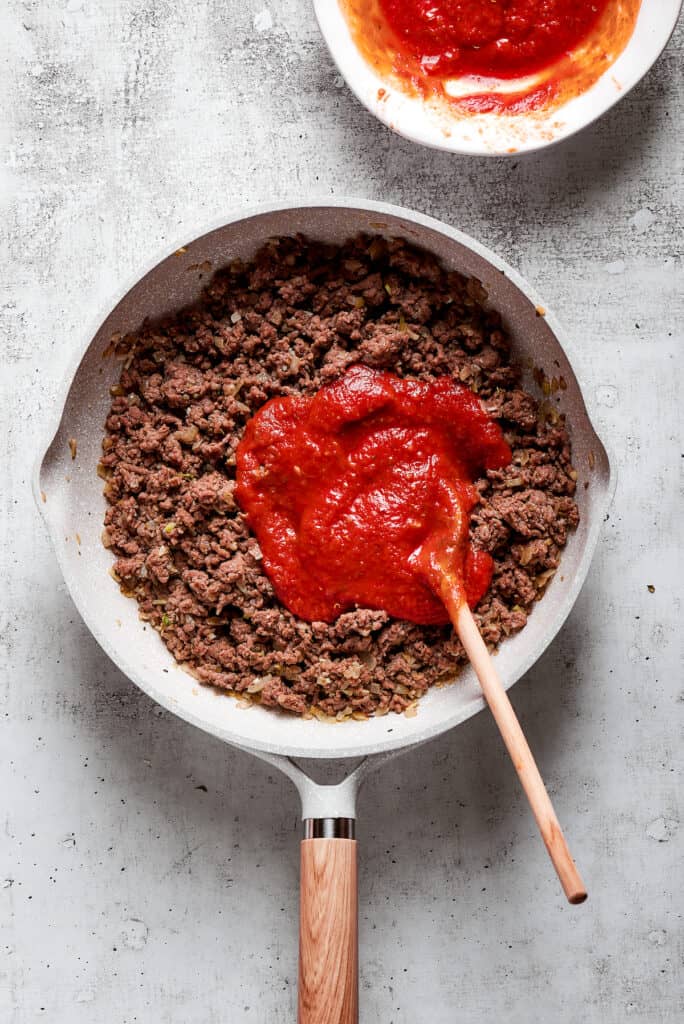Overhead view of marinara sauce on top of cooked ground beef in a skillet with a wooden spoon.