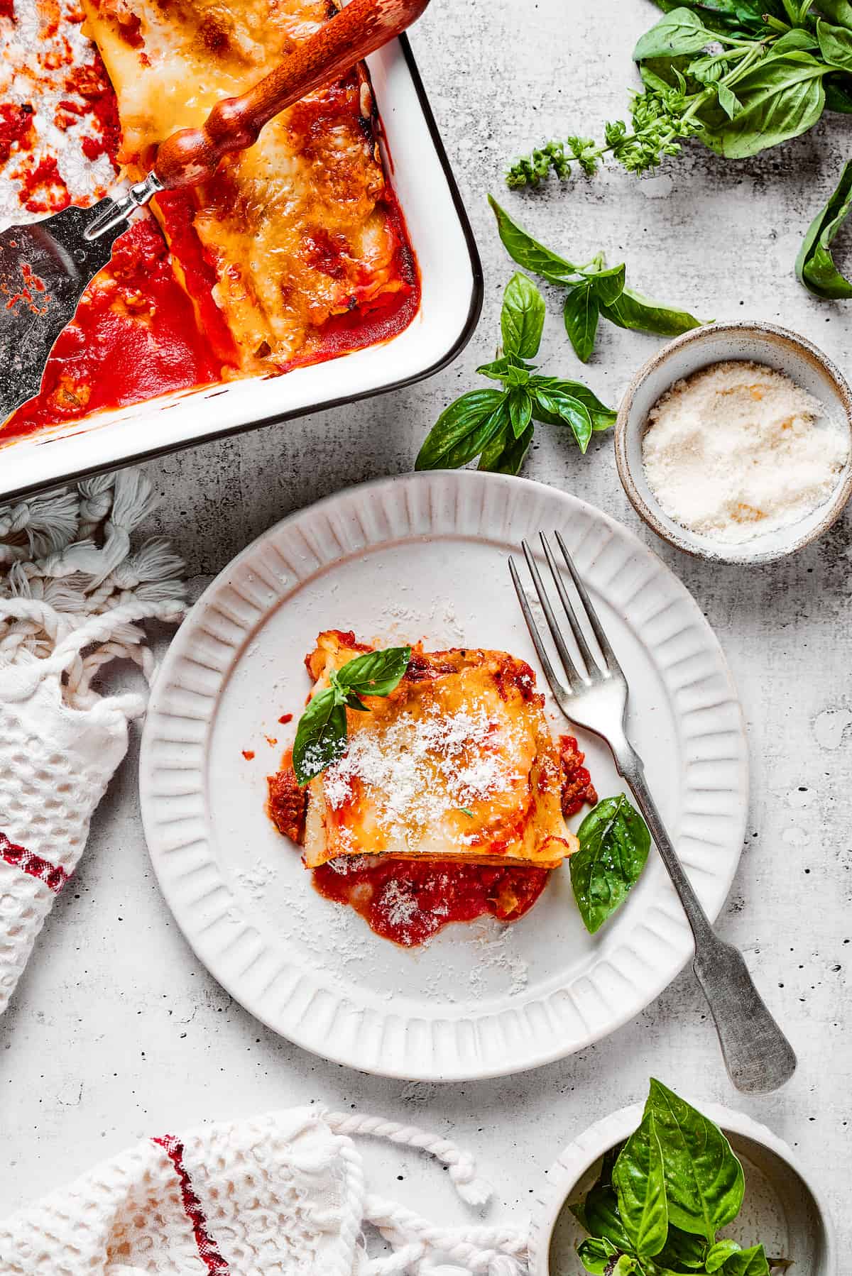 Overhead view of a lasagna roll up on a plate with a fork, next to a baking dish full of them and some basil leaves.