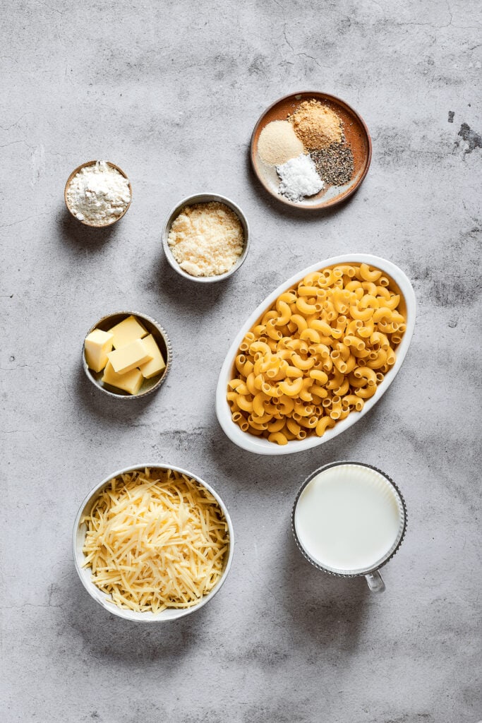 Overhead view of the ingredients needed for mac and cheese: a bowl of elbow macaroni, a bowl of shredded mozzarella cheese, a bowl of grated parmesan, a glass of milk, a bowl of flour, a bowl of butter, and a bowl with salt, pepper, garlic powder, and onion powder.