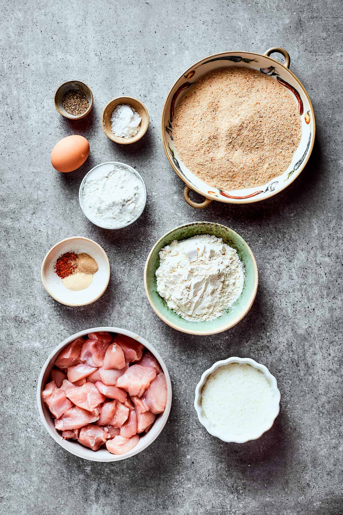 Overhead view of the ingredients needed for bang bang chicken: a bowl of chicken slices, a bowl of flour, a bowl of panko breadcrumbs, a bowl of buttermilk, a bowl of cornstarch, an egg, a bowl of salt, a bowl of pepper, and a bowl of garlic powder, chili powder, and white pepper.