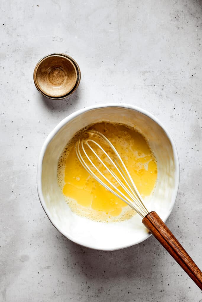 Overhead view of a mixing bowl with eggs, butter, and other wet ingredients mixed together, with a whisk.