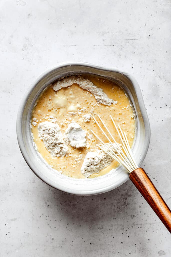 Overhead view of a mixing bowl with wet ingredients, flour, and a whisk.