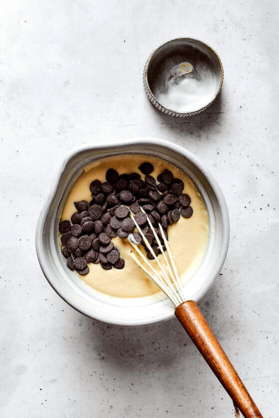 Overhead view of chocolate chips on top of pancake batter in a mixing bowl with a whisk, next to an empty bowl.