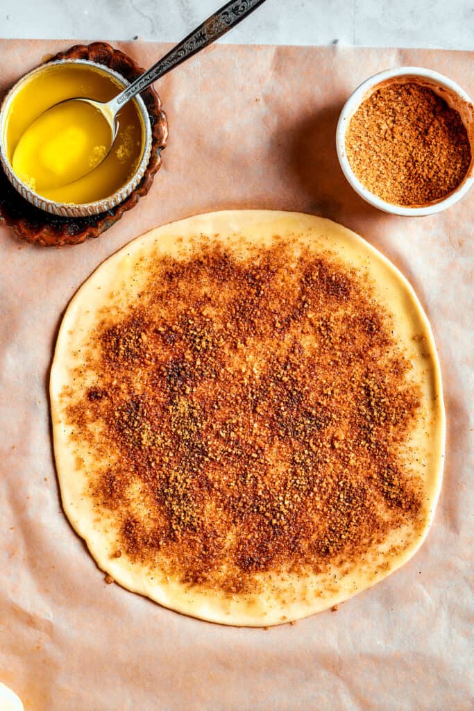 Overhead view of a piece of dough rolled into a circle, covered in cinnamon-sugar, next to a bowl of butter and a bowl of cinnamon-sugar.