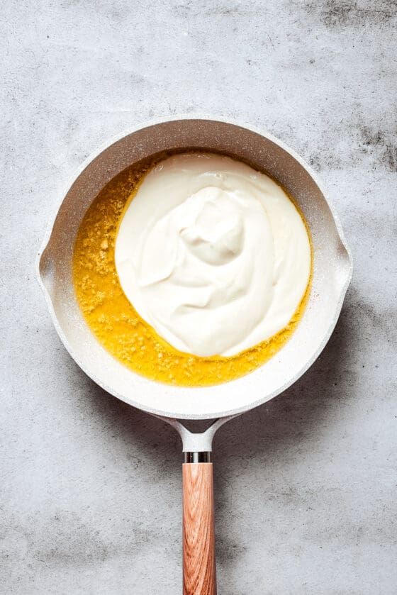Cream cheese and heavy cream sit on top of melted butter with garlic in a white skillet.