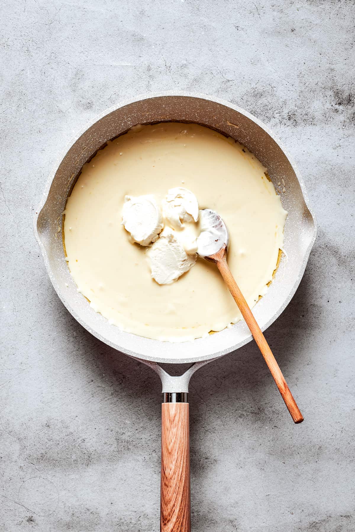 A wooden spoon deftly stirs the cream cheese into the bubbling cream-and-butter mixture in a white skillet, ensuring a perfectly smooth sauce.