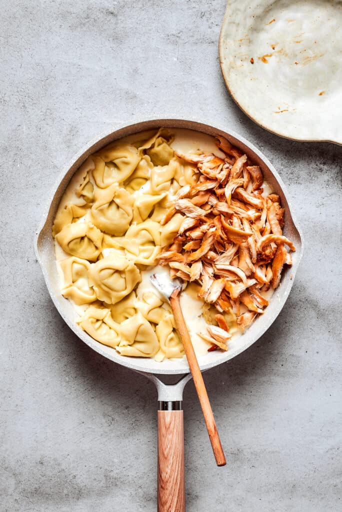 Cheese tortellini and shredded rotisserie chicken are stirred with a wooden spoon into a skillet of cream sauce.