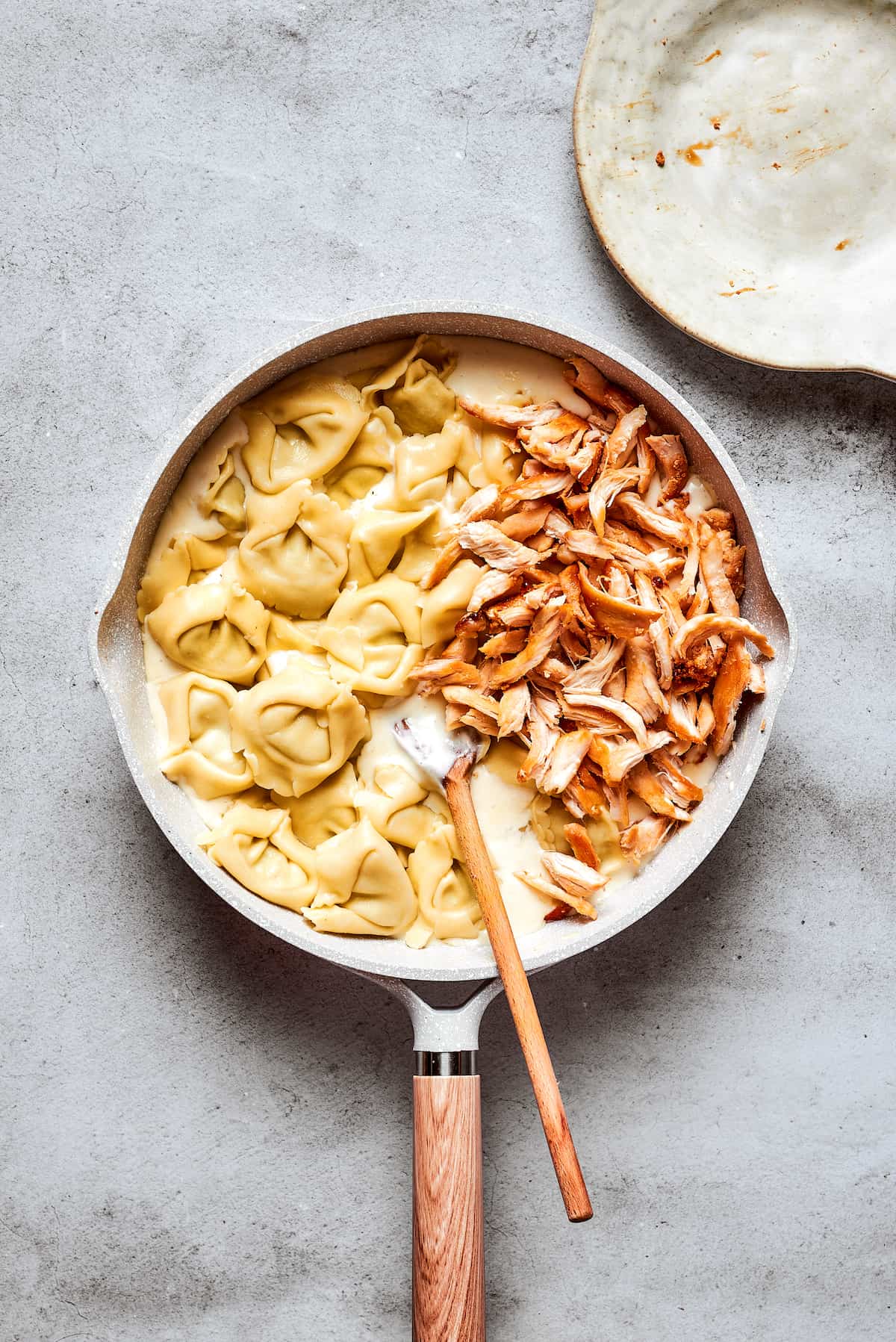 Tender cheese tortellini and flavorful shredded rotisserie chicken are gently folded into the luscious cream sauce in a white skillet with a wooden spoon, ready to be coated in cheesy goodness.