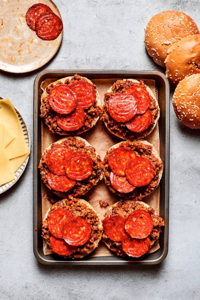Pepperoni is placed on top of pizza burgers on a tray.