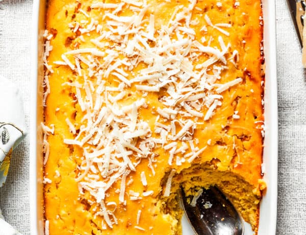 A white baking pan of baked corn souffle with a piece cut out and a spoon in the lower right side.