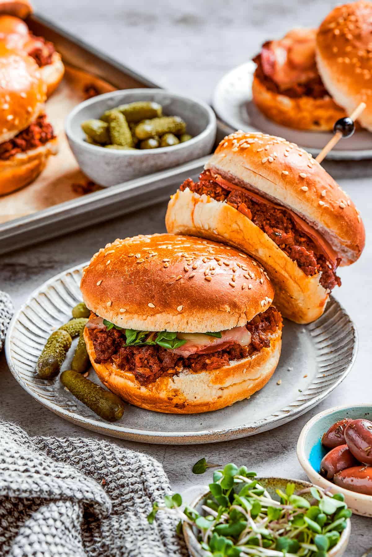 Two pizza burgers served on a plate with a few other burgers and pickles in the background.