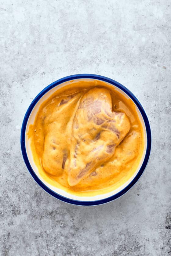 Chicken breasts marinate in a white bowl of yellow honey mustard sauce.