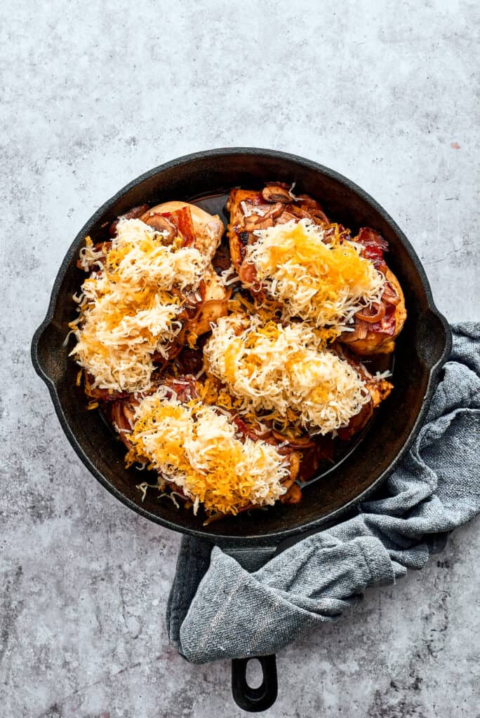 Four chicken breasts are topped with bacon and cheese in a cast iron skillet.