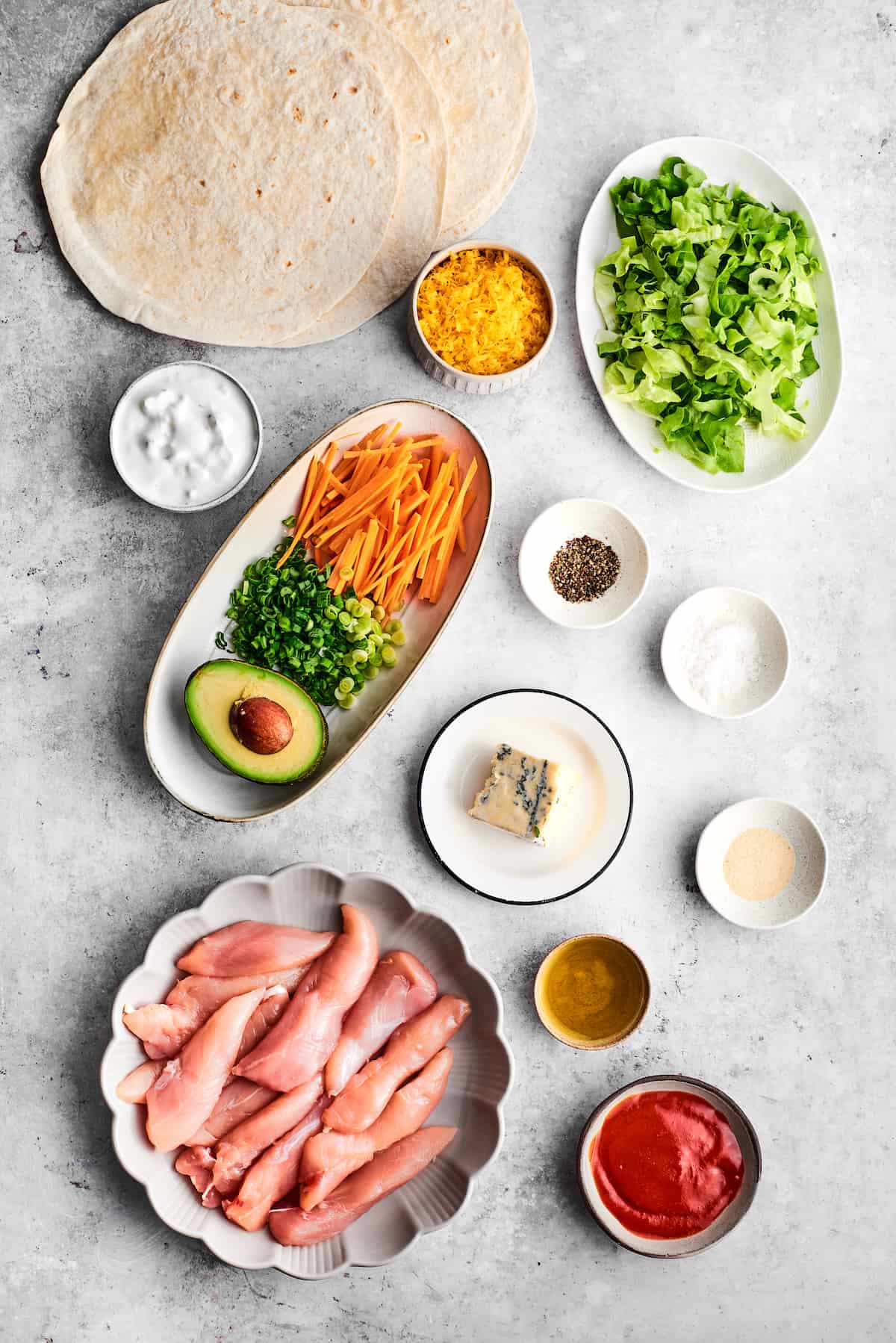 The ingredients for buffalo chicken wrap are shown portioned out: chicken tenders, olive oil, salt and pepper, garlic powder, hot sauce, ranch dressing, avocado green onions, carrots, tortillas, cheddar cheese, and blue cheese.