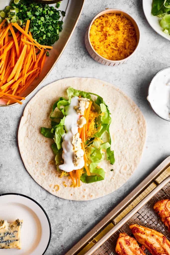 Ranch dressing is drizzled over a tortilla and vegetables to make a buffalo chicken wrap.