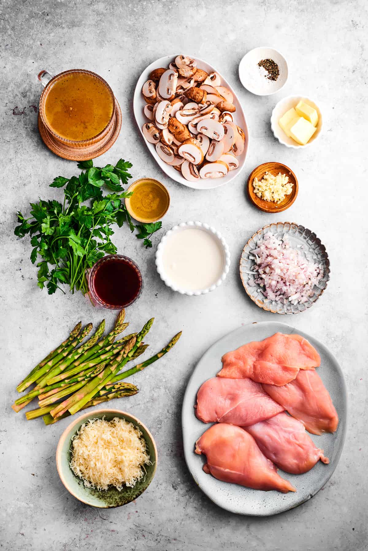 The ingredients for chicken madeira are shown portioned out: chicken, mushrooms, asparagus, parsley, salt and pepper, butter, olive oil, madeira, garlic, mozzarella, cream, broth.