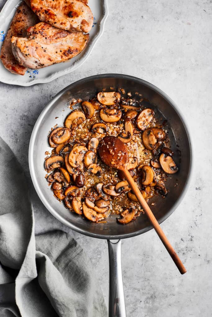 A wooden spoon stirs a metal skillet full of cooked sliced mushrooms.