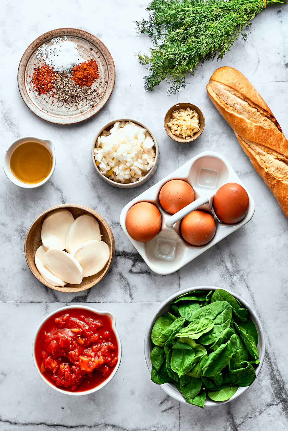 The ingredients for eggs in purgatory are shown on a white background: eggs, bread, fresh mozzarella cheese, spinach, diced tomatoes, garlic, olive oil, spices, dill.