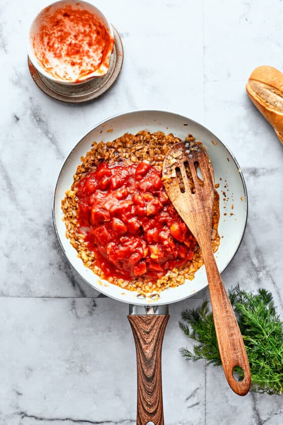 Thick diced tomatoes are stirred into a white skillet with a wooden spoon.