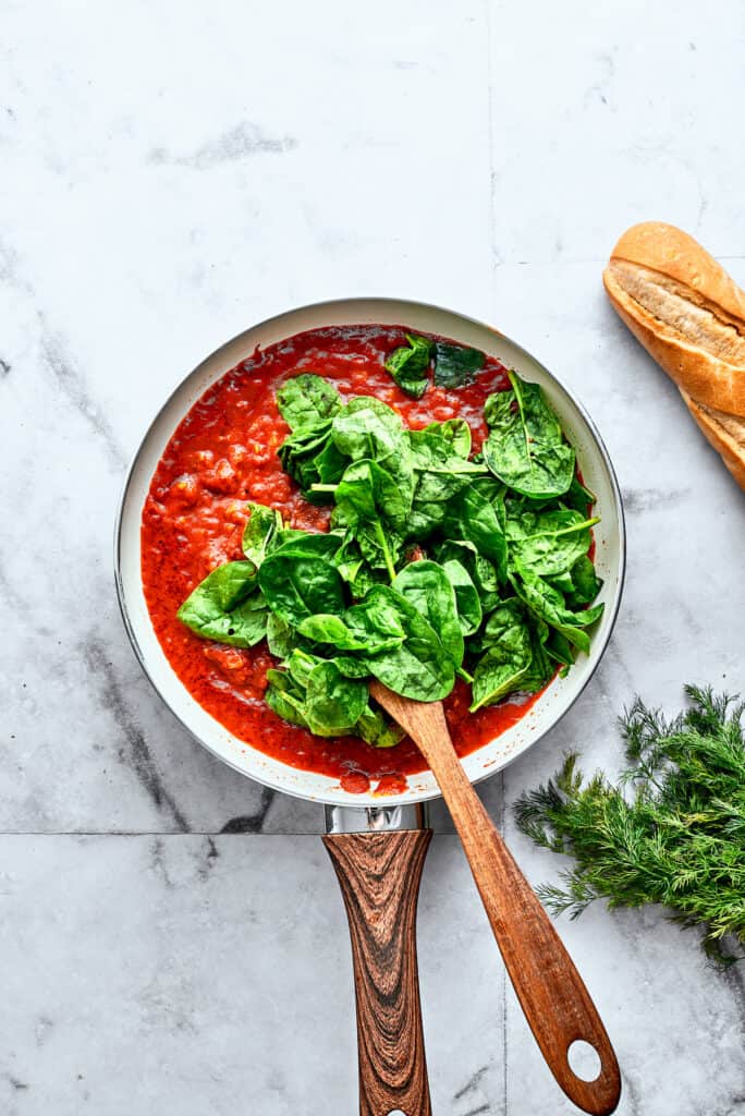 Bright green spinach leaves are stirred into a skillet of homemade tomato sauce.