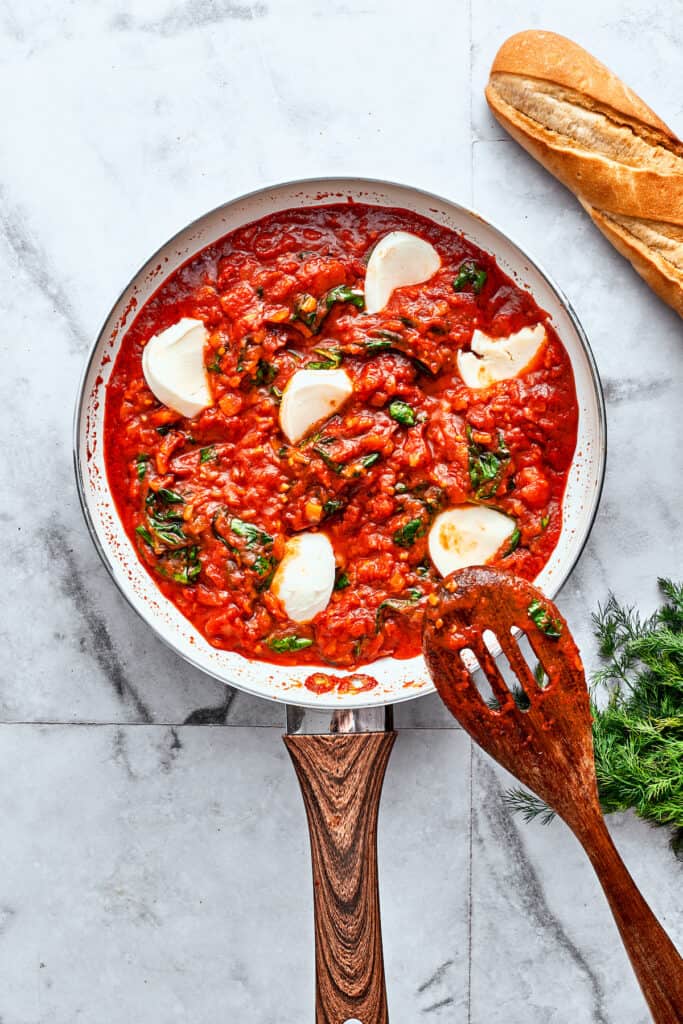 Slices of fresh mozzarella cheese are seen peeking through a hearty red sauce in a skillet with a wooden spoon.