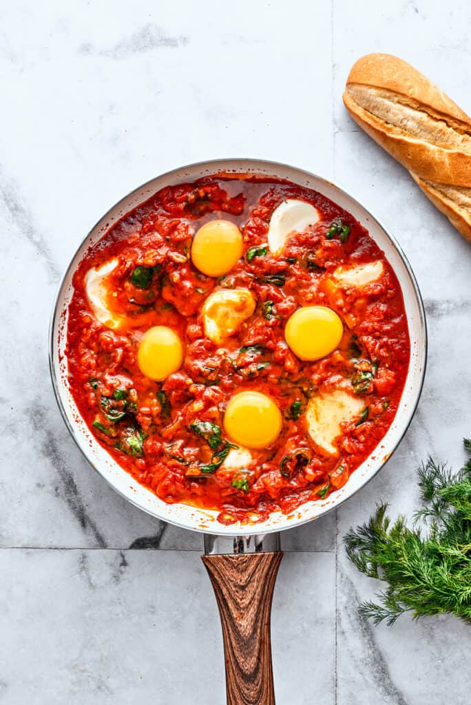 A skillet of eggs in purgatory shows white slices of mozzarella and yellow egg yolks in red sauce.