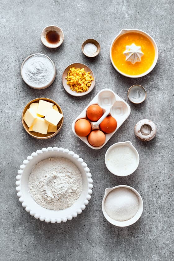 The ingredients needed for orange cake are shown portioned out: eggs, flour, sugar, orange juice, orange zest, orange liqueur, butter, buttermilk, baking powder, salt.