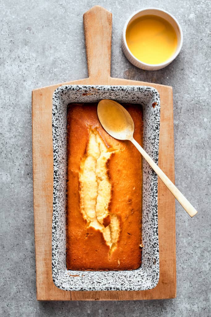 The baked loaf pan of orange cake is shown with a spoon resting on it and a bowl of orange glaze next to it ready to be added to the top.