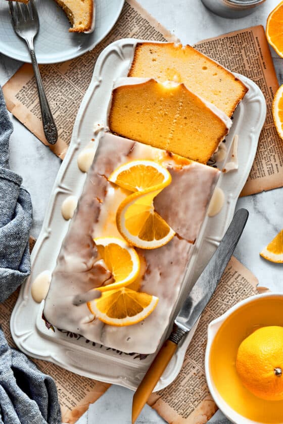 A sliced orange loaf cake topped with orange glaze, orange slices, and wish more oranges surrounding it.