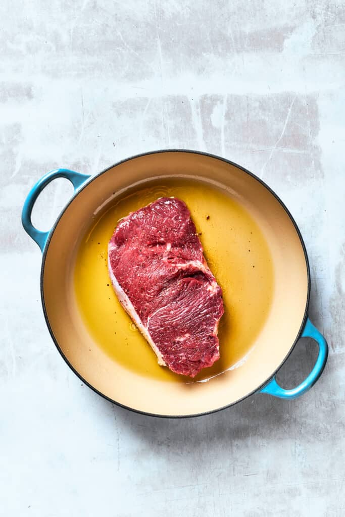 A piece of steak cooks in olive oil in a pan.