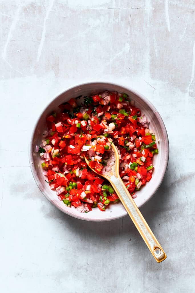 A bowl of Pico de Gallo is mixed with a wooden spoon.
