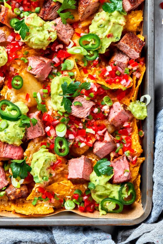 A corner of a pan of steak nachos is shown colorfully topped with guacamole and tomatoes.