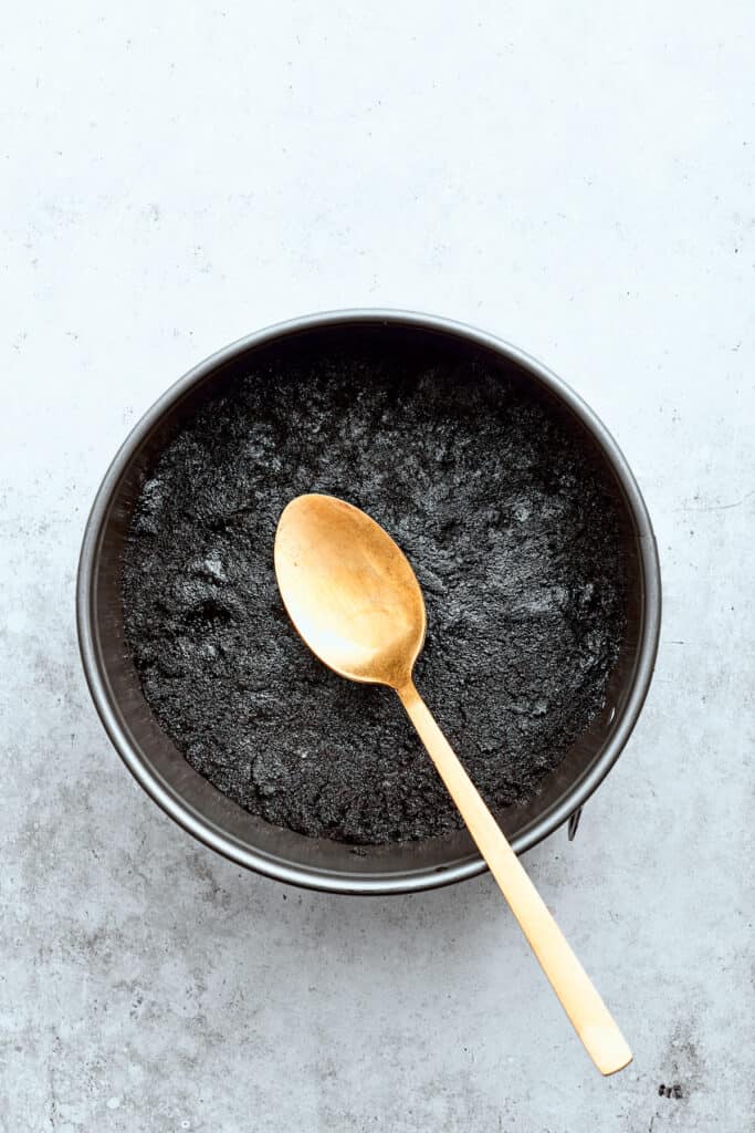 A spoon pats a chocolate cookie crust into a springform baking pan.