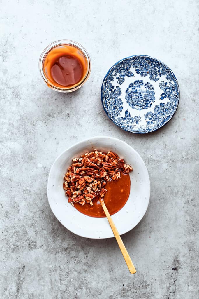 Pecans and caramel sauce are stirred together with a wooden spoon in a white bowl.
