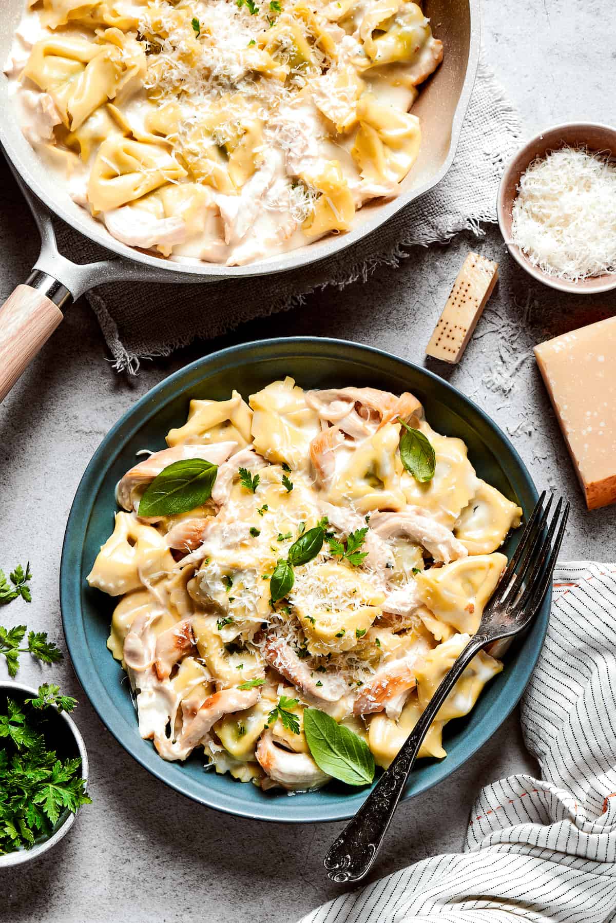 A blue bowl of chicken tortellini alfredo with a fork balanced on the edge of the bowl and a skillet of chicken tortellini placed near it.