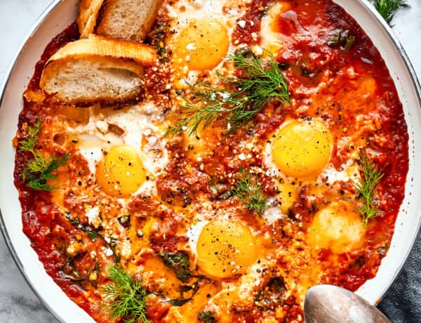 Cooked eggs in purgatory in a skillet are garnished with parsley and toasted bread with a silver spoon resting on its edge.