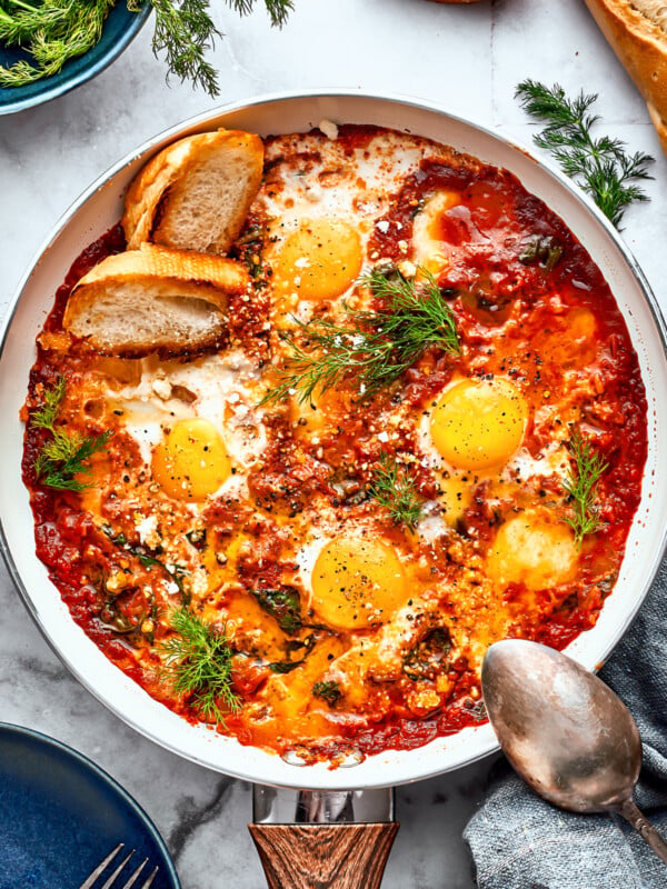 Cooked eggs in purgatory in a skillet are garnished with parsley and toasted bread with a silver spoon resting on its edge.