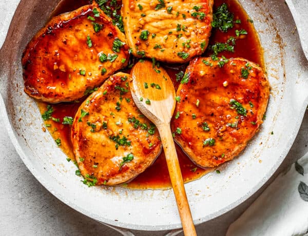 Four honey garlic pork chops cooking in a skillet with a wooden spoon stirring through.