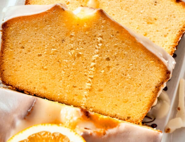 Two slices of glazed orange cake rest atop each other, with the rest of the loaf visible nearby topped with orange slices.